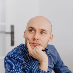 Bald man in blue shirt thoughtfully holding a pen indoors, gazing into distance.